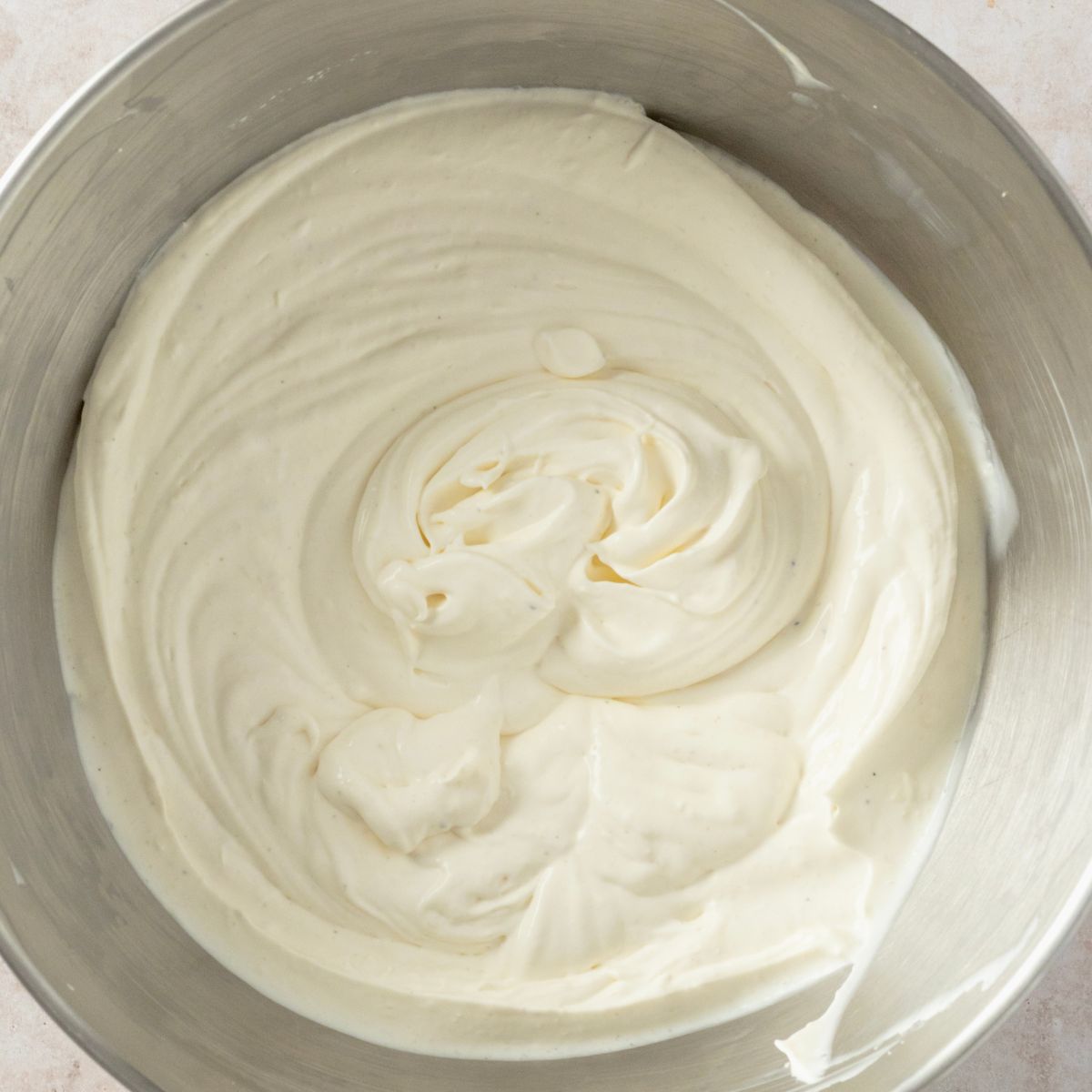 Overhead view of thick, freshly whipped cream with soft swirls in a stainless steel mixing bowl.