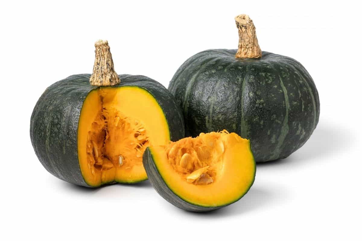 Whole and cut kabocha squash with dark green skin and bright orange flesh on a white background.