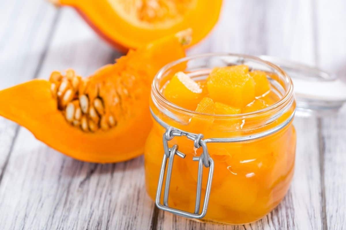 Glass jar filled with preserved pumpkin cubes, with fresh pumpkin slices in the background.