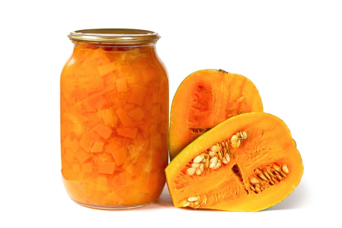 Jar of preserved pumpkin beside fresh pumpkin wedges on a white background.