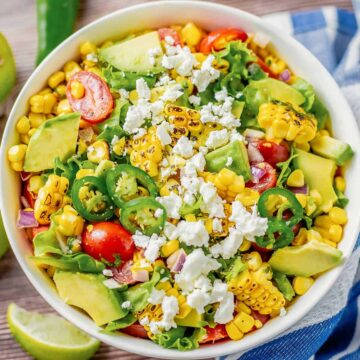 grilled corn salad with lettuce, avocado, tomatoes, onion in a white bowl on a wooden table with blue white striped towel next to it