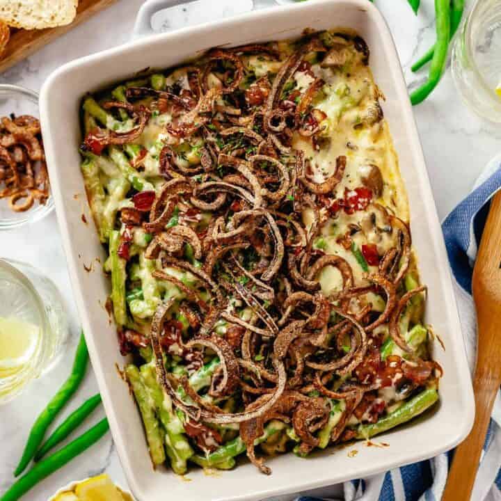 Overhead shot of a baked green bean casserole with bacon on a marble table with lemon slices, scattered green beans, bread and crispy red onions.