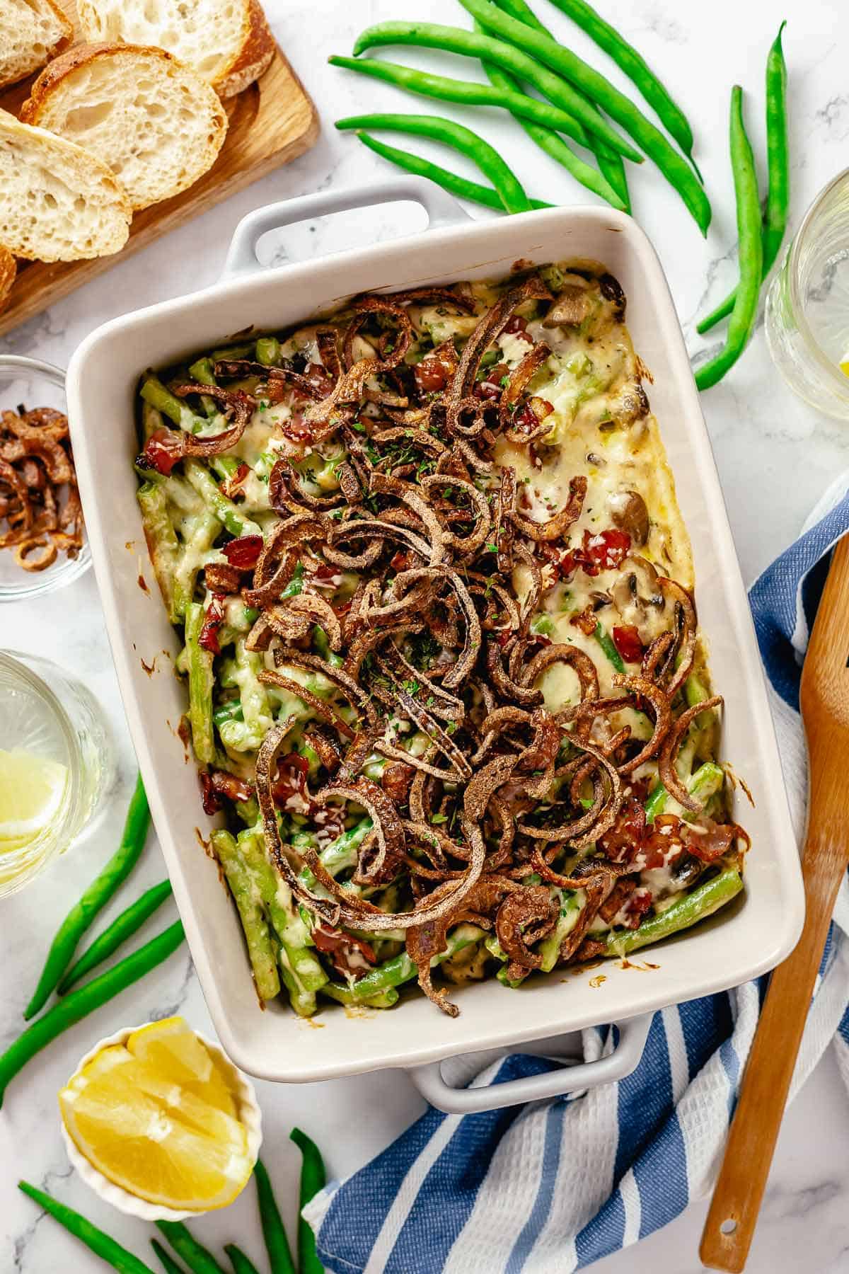 Overhead shot of a baked green bean casserole with bacon on a marble table with lemon slices, scattered green beans, bread and crispy red onions.