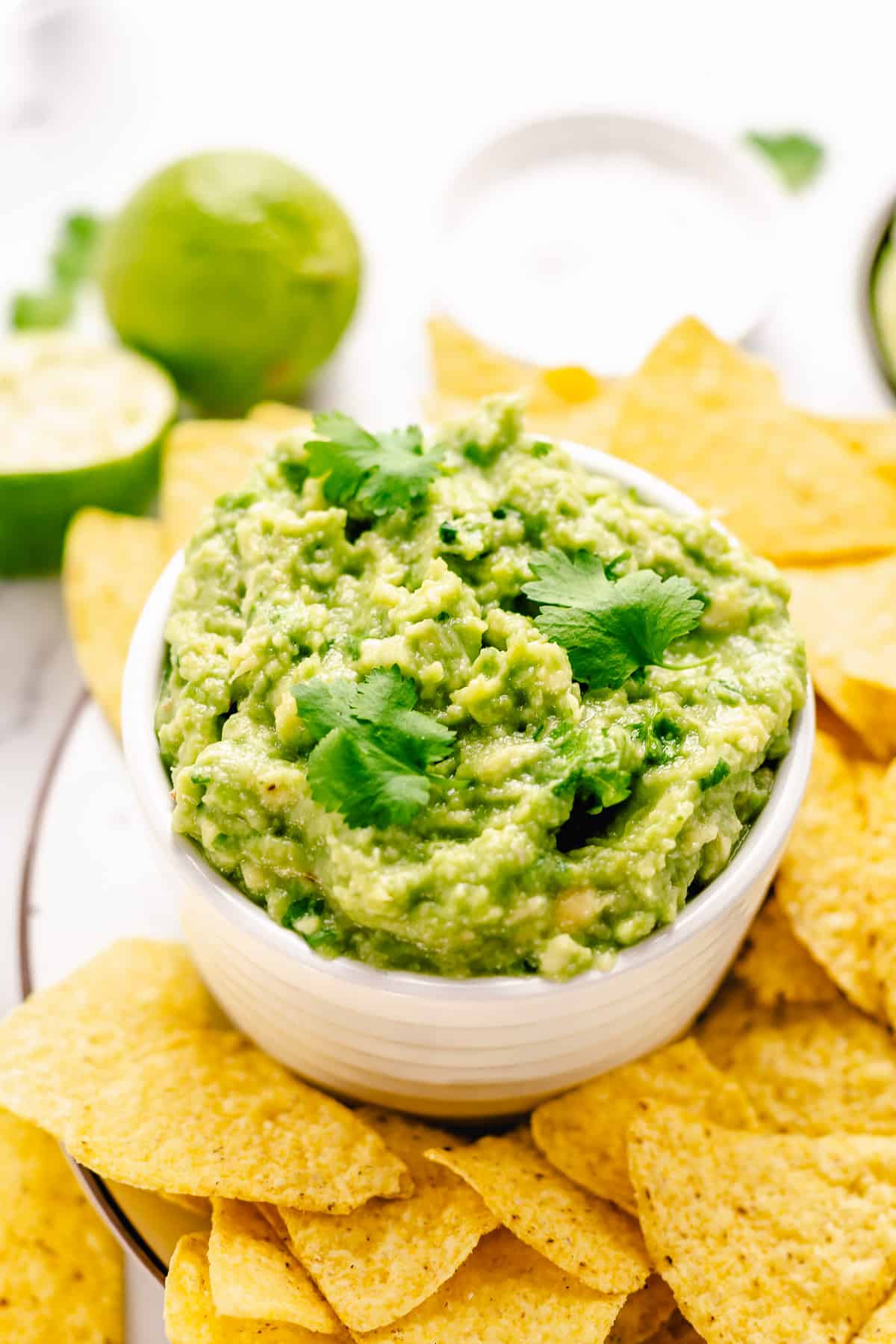 Simple guacamole in a white bowl with nacho chips around on a white marble background