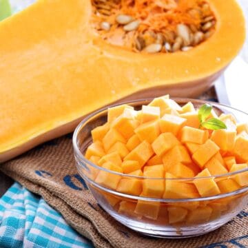 Halved butternut squash with seeds and a glass bowl of peeled, cubed butternut squash on a kitchen surface.