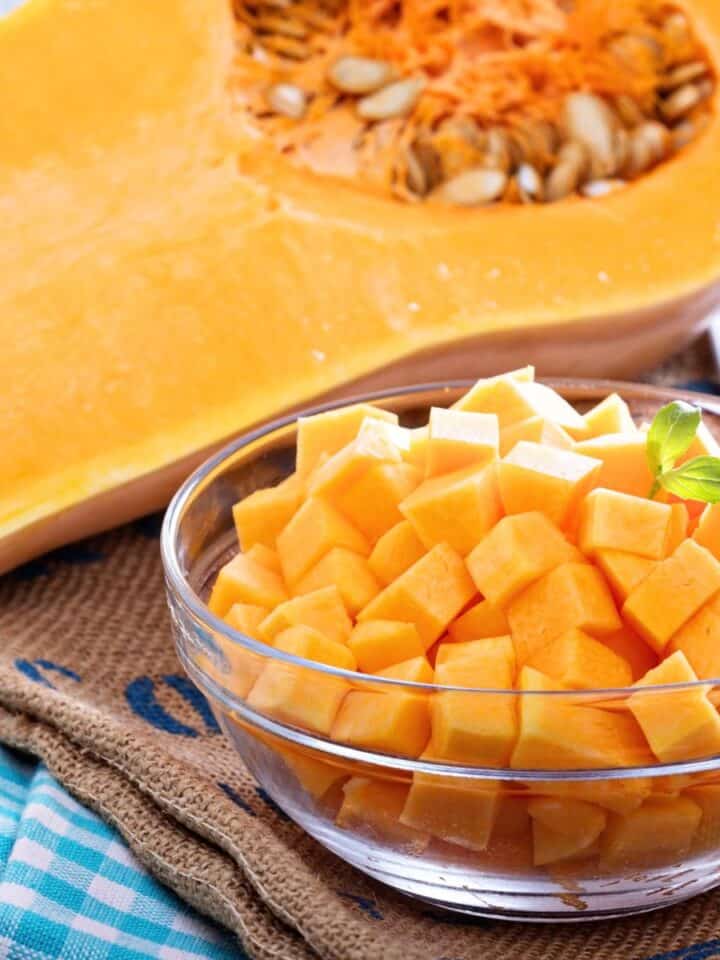 Halved butternut squash with seeds and a glass bowl of peeled, cubed butternut squash on a kitchen surface.
