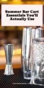 Close-up of stainless steel bar tools, including a cocktail shaker, double jigger, mixing glass, and bar spoon, arranged on a bar mat with warm, blurred lights in the background.