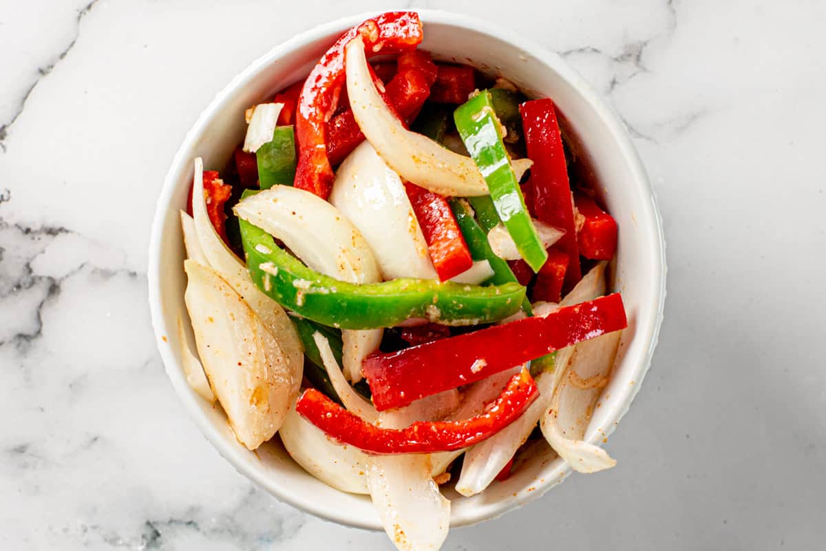 onions and bell pepper strips marinating in a bowl with fajita seasonings