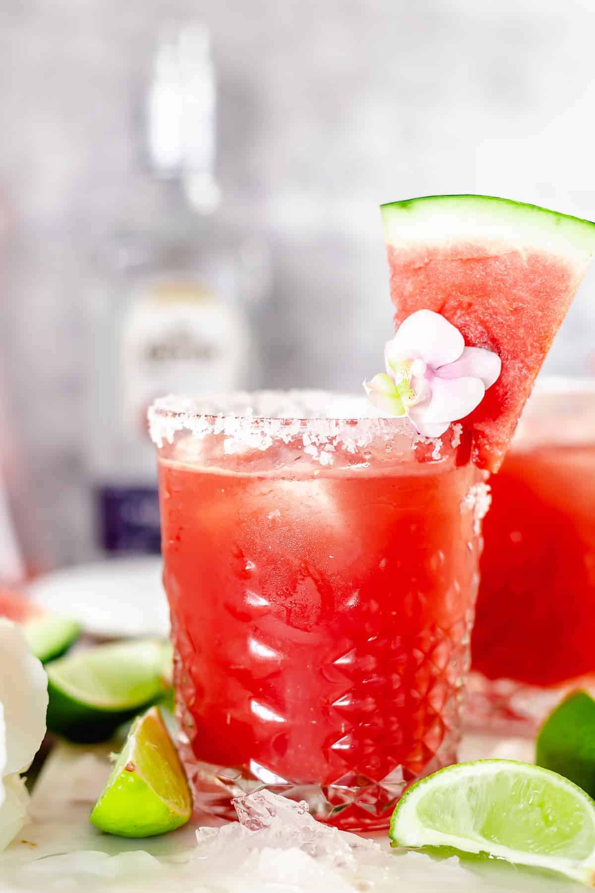 angle view shot of watermelon margarita in whisky glass garnished with purple pink flower, and watermelon slice