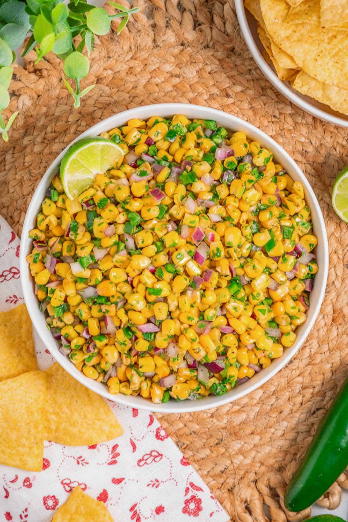 Chipotle-style corn salsa in a bowl with lime wedges and tortilla chips on a neutral background.