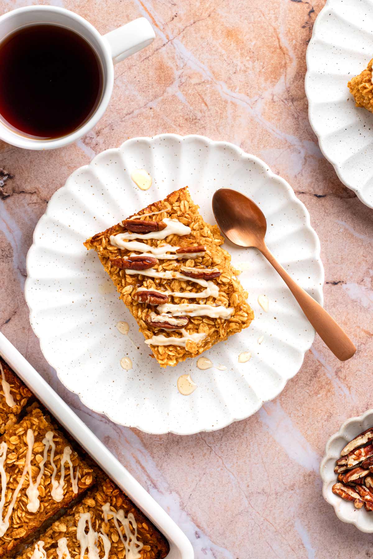 One square of glazed pumpkin baked oatmeal topped with pecans on a white scalloped plate with a copper spoon and a cup of coffee.