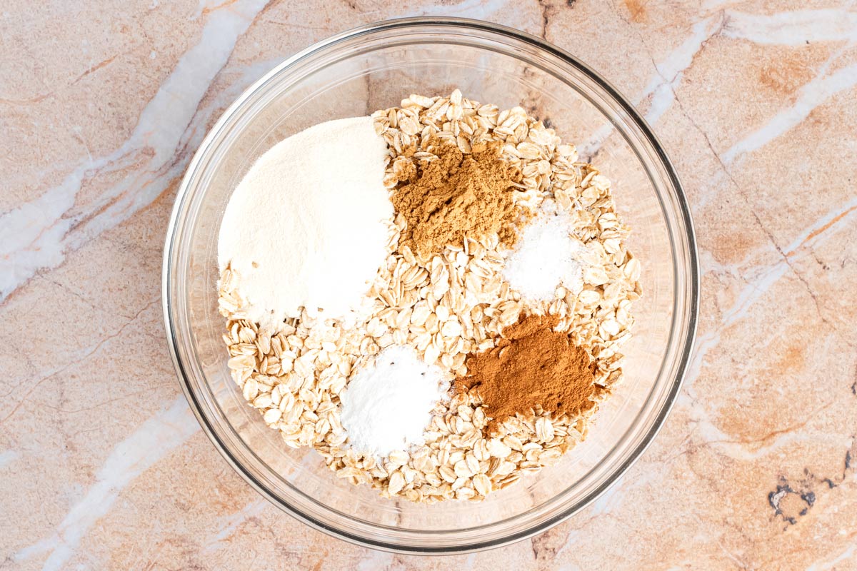 Glass bowl of dry ingredients for pumpkin baked oatmeal: rolled oats, protein powder, pumpkin pie spice, cinnamon, baking powder, and salt.