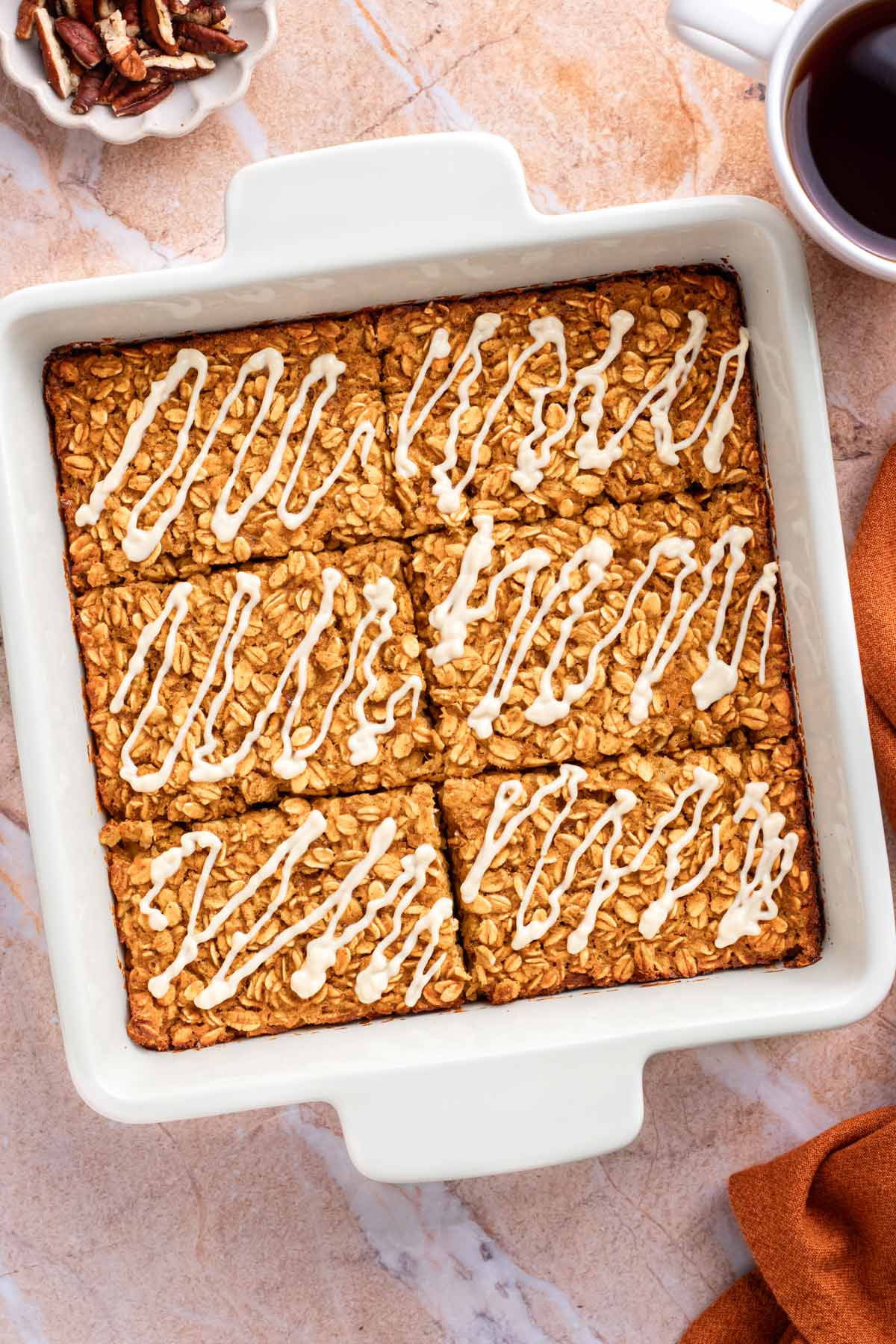 Pumpkin baked oatmeal squares drizzled with white cream cheese glaze in a white baking dish, next to a bowl of pecans and a cup of coffee.