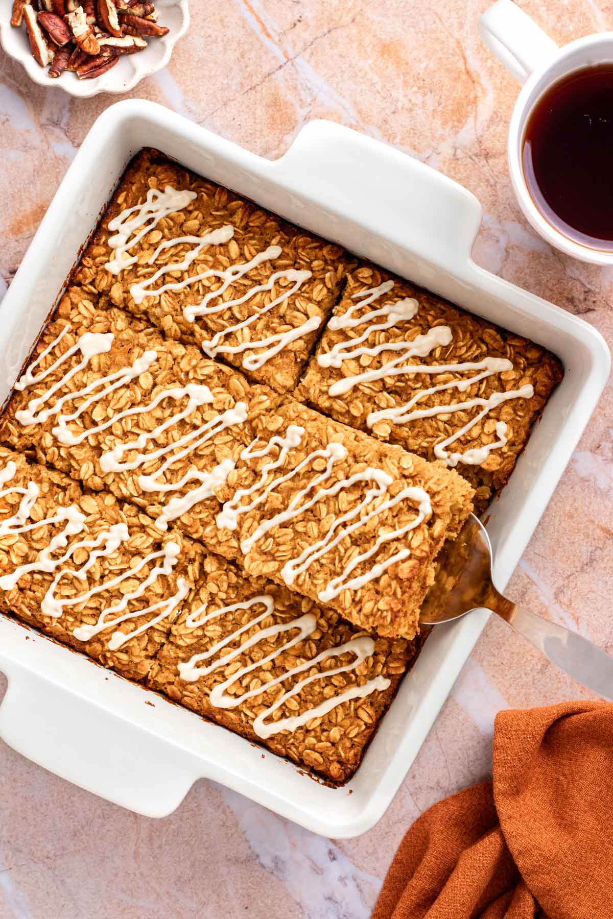 Overhead view of pumpkin baked oatmeal drizzled with cream cheese glaze in a white square baking dish, with a cup of coffee, a bowl of pecans, and one square lifted out with a spatula.
