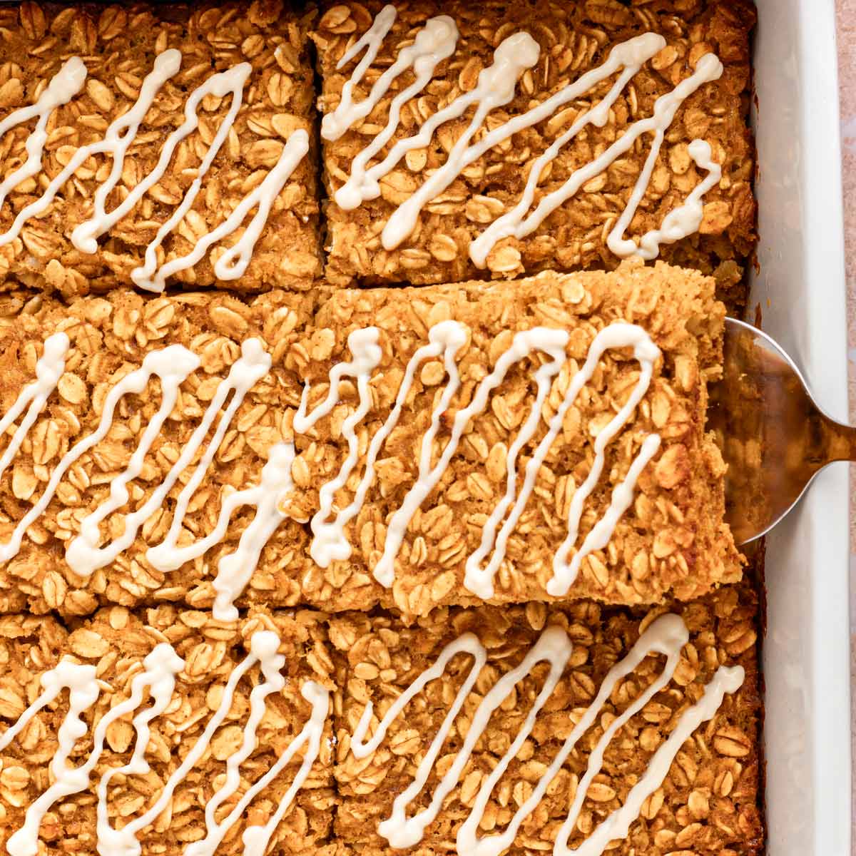 Close up of glazed pumpkin baked oatmeal squares with a spatula lifting one out of the white baking dish.